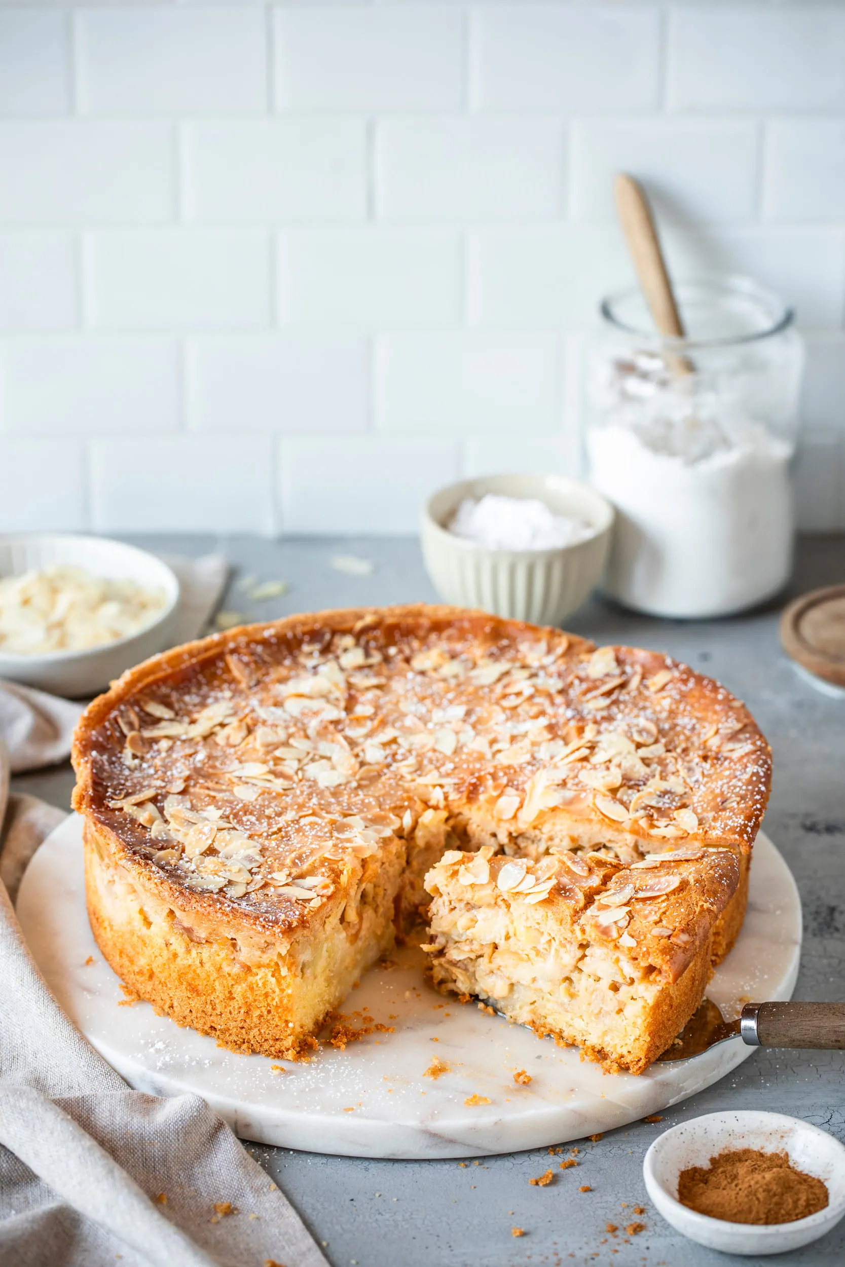 Slice of almond-topped apple cake on a white marble serving board with a cake server and baking ingredients in the background.