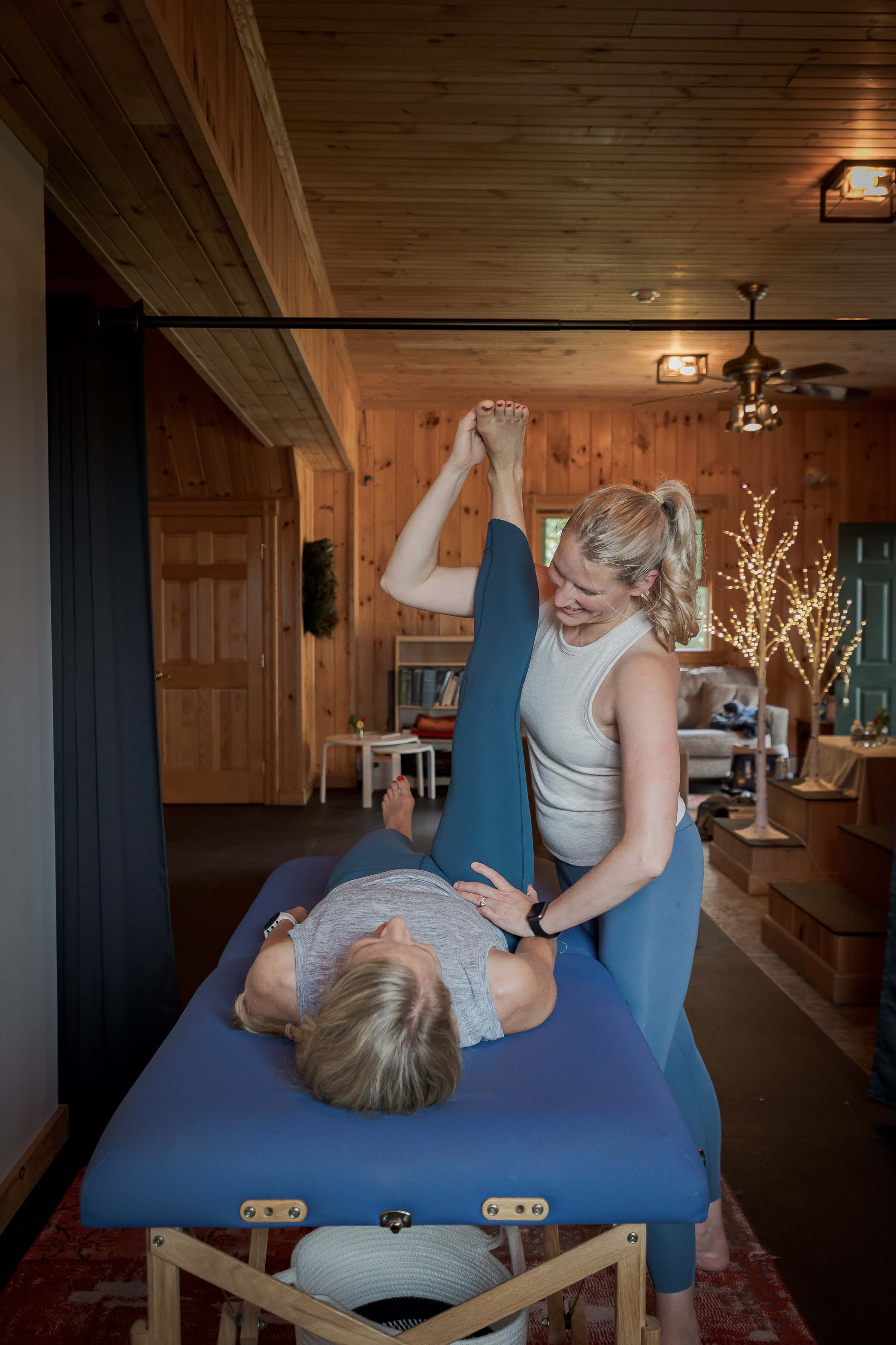 A woman assists another woman lying on a massage table by stretching her raised leg in a wooden room.