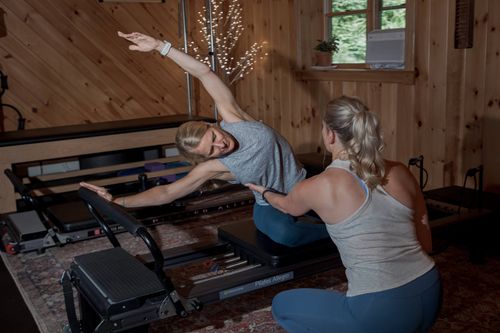 Two women in a wooden-paneled room practicing Pilates on a reformer machine, one instructing the other in a side stretch pose. stowe-physical-therapy-pilates