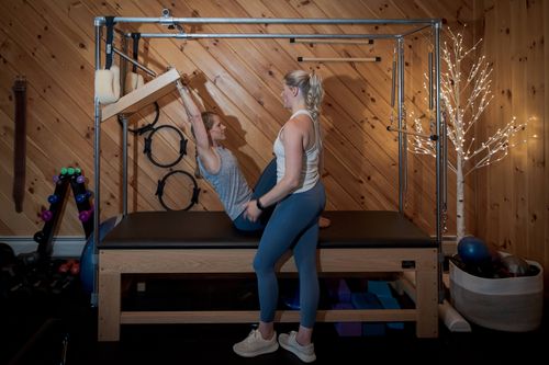 Two women in workout clothes using a Pilates reformer machine in a wood-paneled room with fitness equipment and a lit decorative tree.