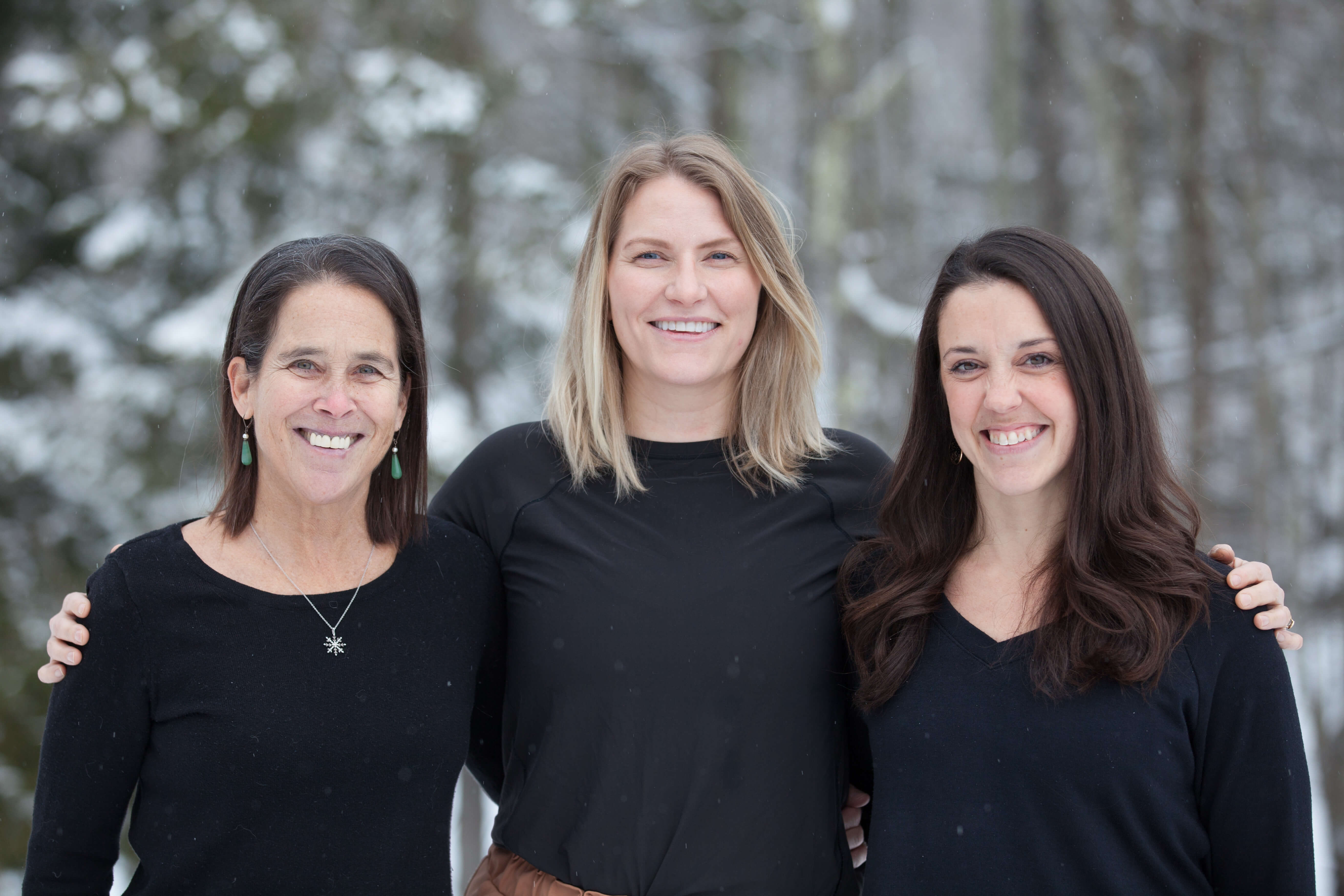 Three smiling women wearing black standing outdoors with a snowy forest background.