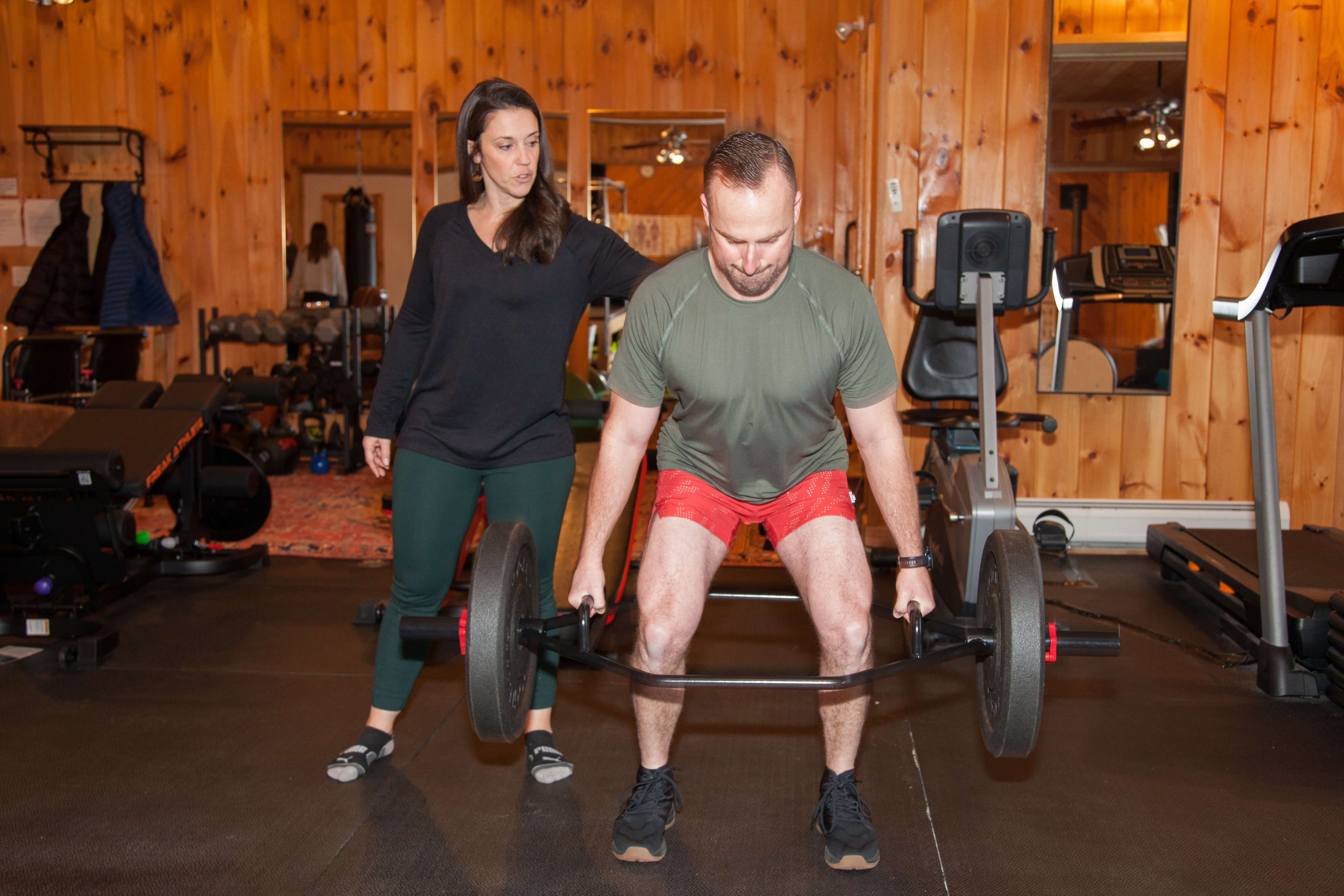Man lifting a barbell with weights while a woman coaches him in a wood-paneled gym.