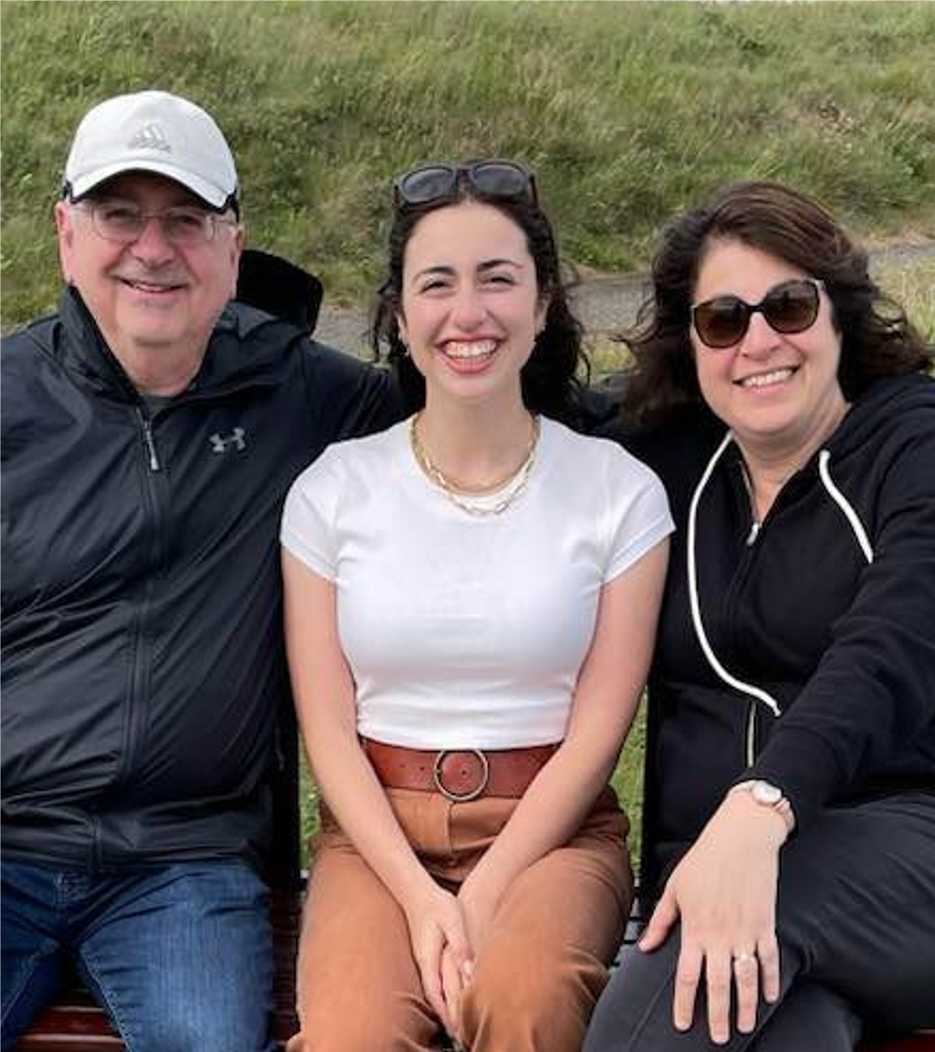 Three people sitting outdoors on a bench, smiling; a man in a black jacket and white cap, a woman in a white t-shirt and brown pants, and a woman in a black hoodie and sunglasses.