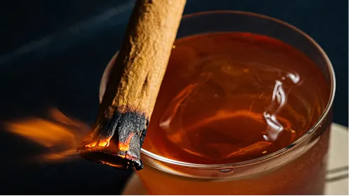 Close-up of a lit cinnamon stick resting on the rim of a glass filled with amber liquid.