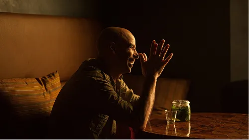 Man smiling with hands clasped near face, sitting at a table with a jar and glass in dim lighting.
