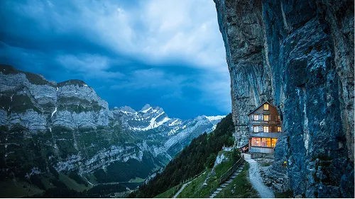 Mountain lodge built into a cliffside illuminated at dusk, overlooking rugged mountain peaks under a cloudy sky.