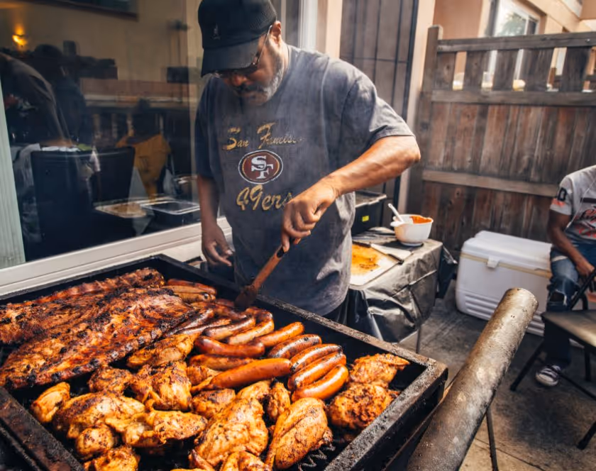 Man wearing a San Francisco 49ers shirt grilling assorted meats including sausages, chicken wings, and ribs outdoors.