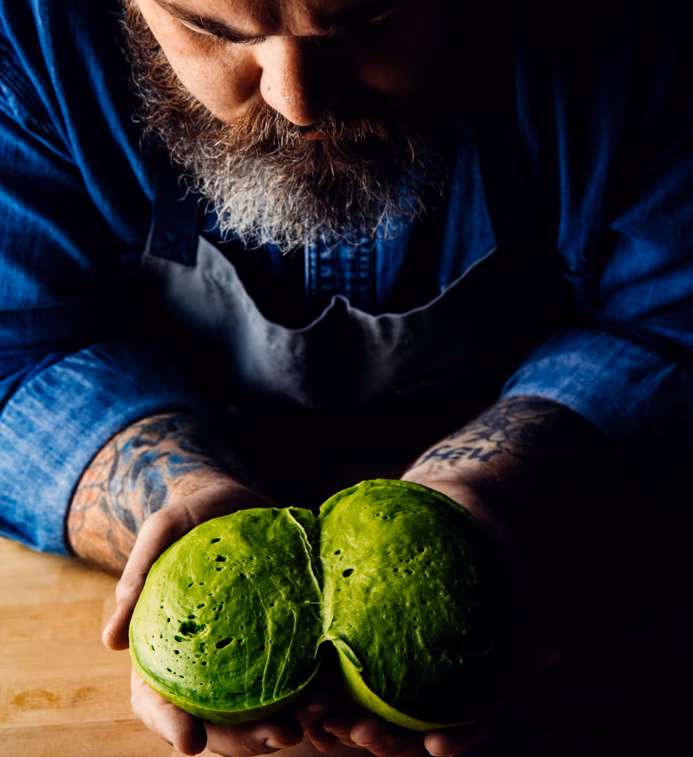 Bearded man with tattoos holding two pieces of green spinach bread dough in his hands over a wooden surface.