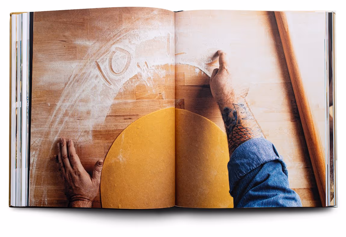 Open cookbook showing overhead view of hands preparing dough on a wooden surface dusted with flour next to a rolling pin.