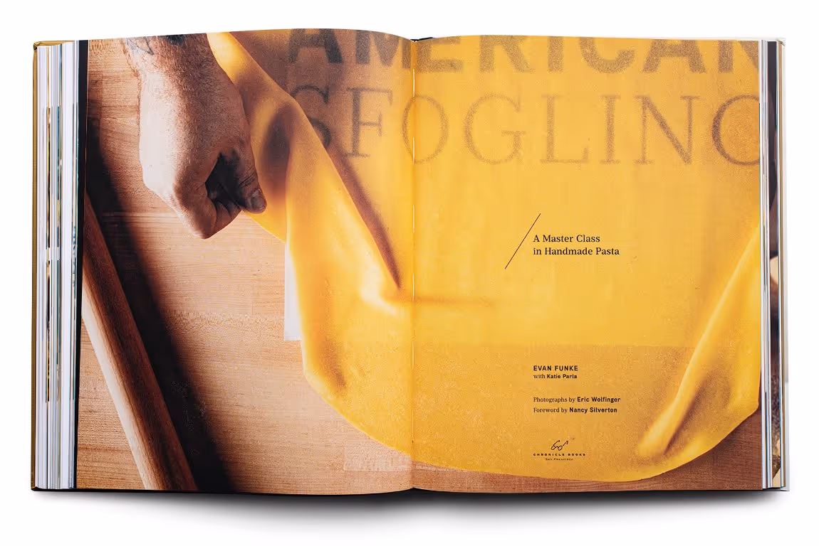 Open cookbook showing a person's hand lifting a sheet of yellow pasta dough on a wooden surface with text about a handmade pasta master class.
