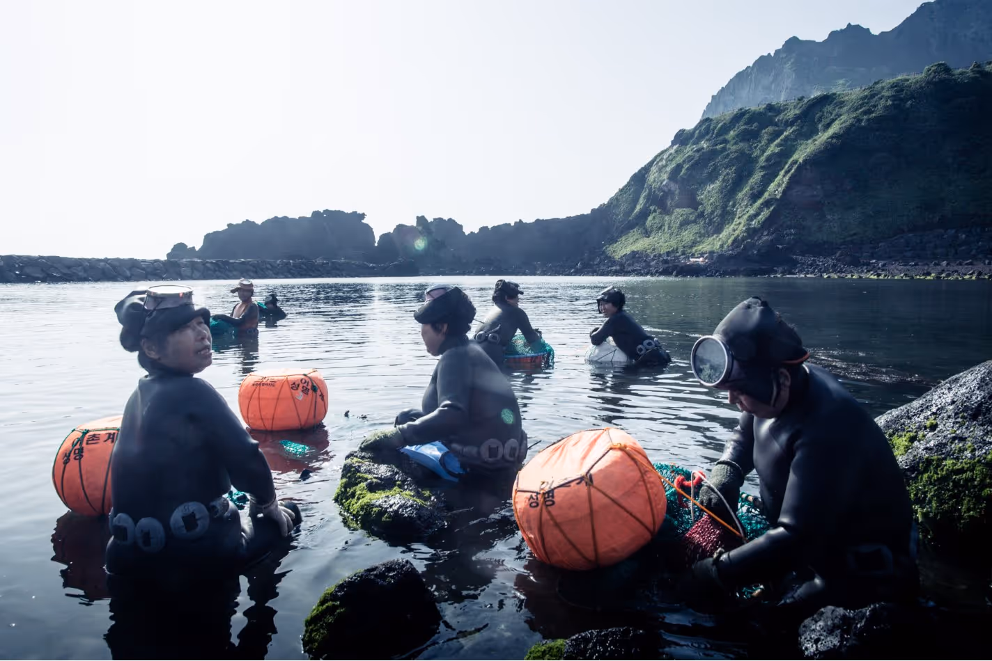 Female divers in black wetsuits preparing for sea diving with orange buoys in a calm coastal area with rocky hills.