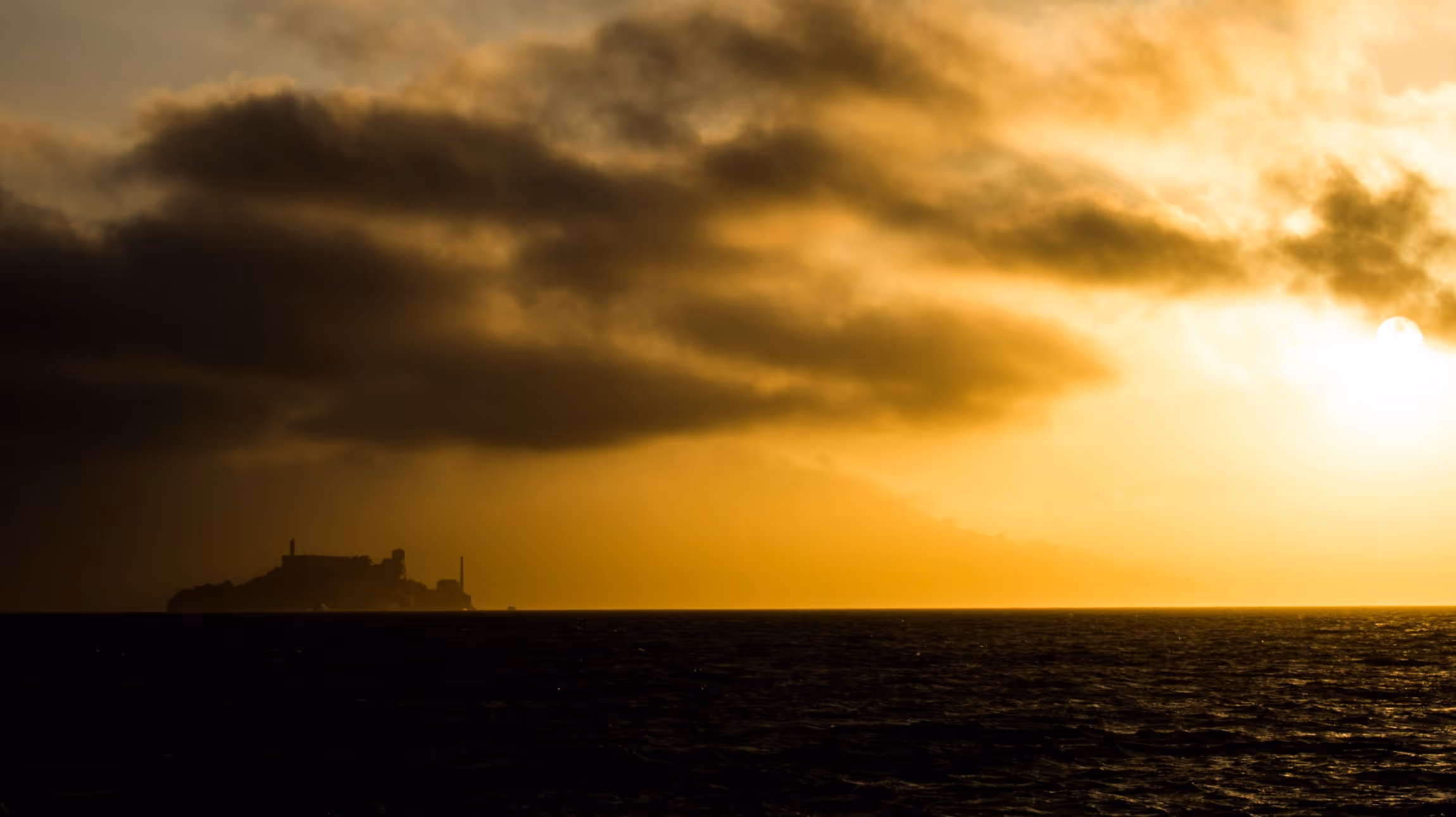 Sunset casting golden light over dark ocean with a silhouetted island and scattered clouds.