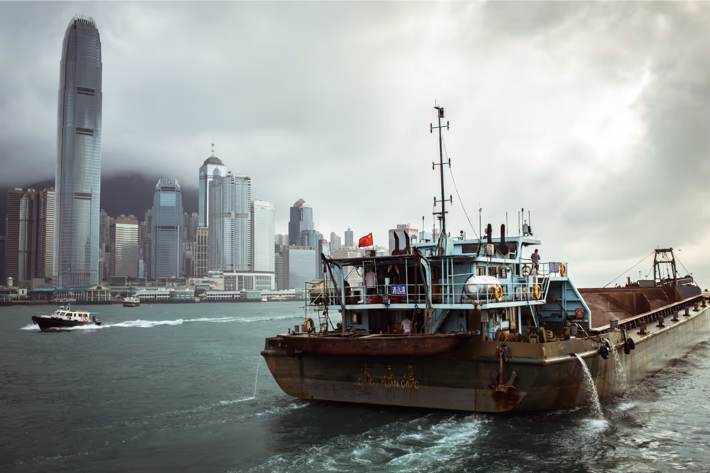 Large cargo ship with Chinese flag sailing in harbor with city skyscrapers and cloudy sky in the background.
