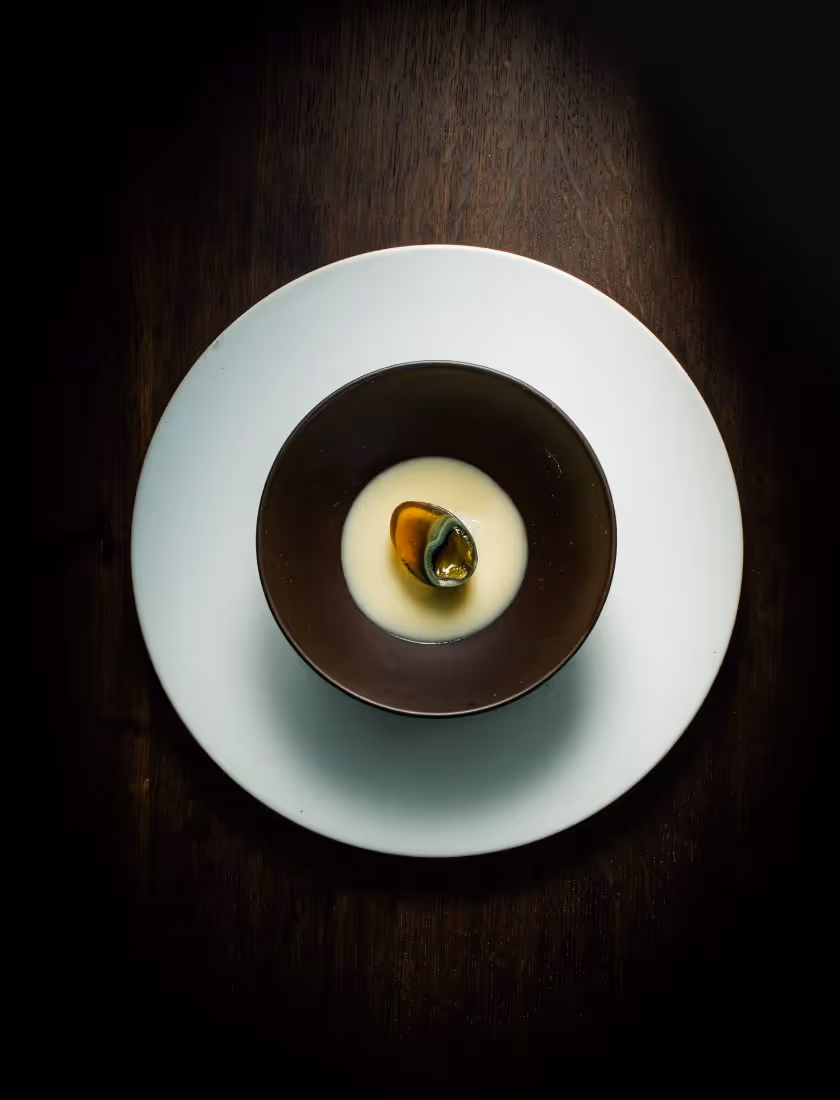Small dark bowl with creamy soup and a half clam shell on a white plate over a dark wooden surface.