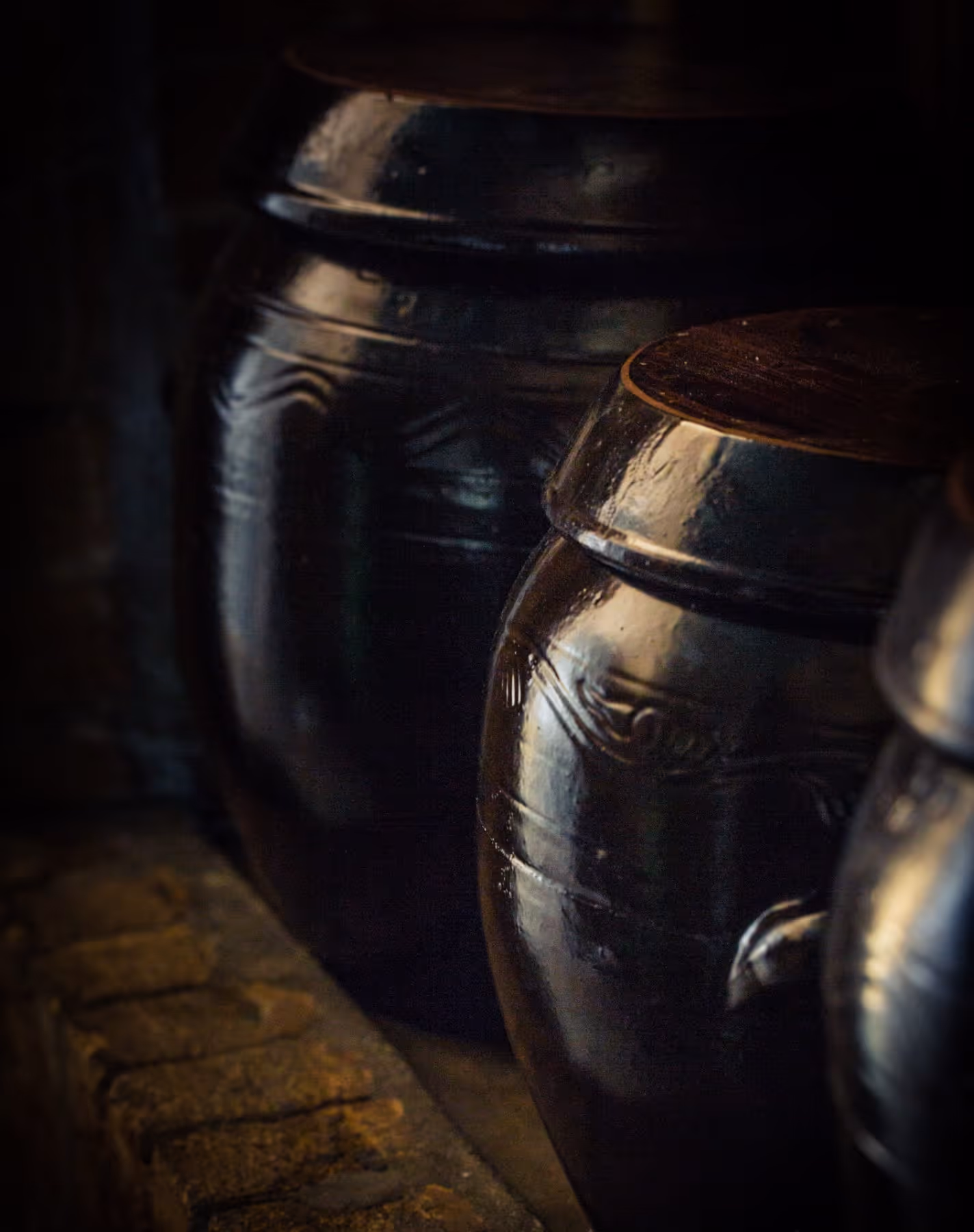 Close-up of traditional dark brown clay jars with lids placed on a stone surface in dim lighting.