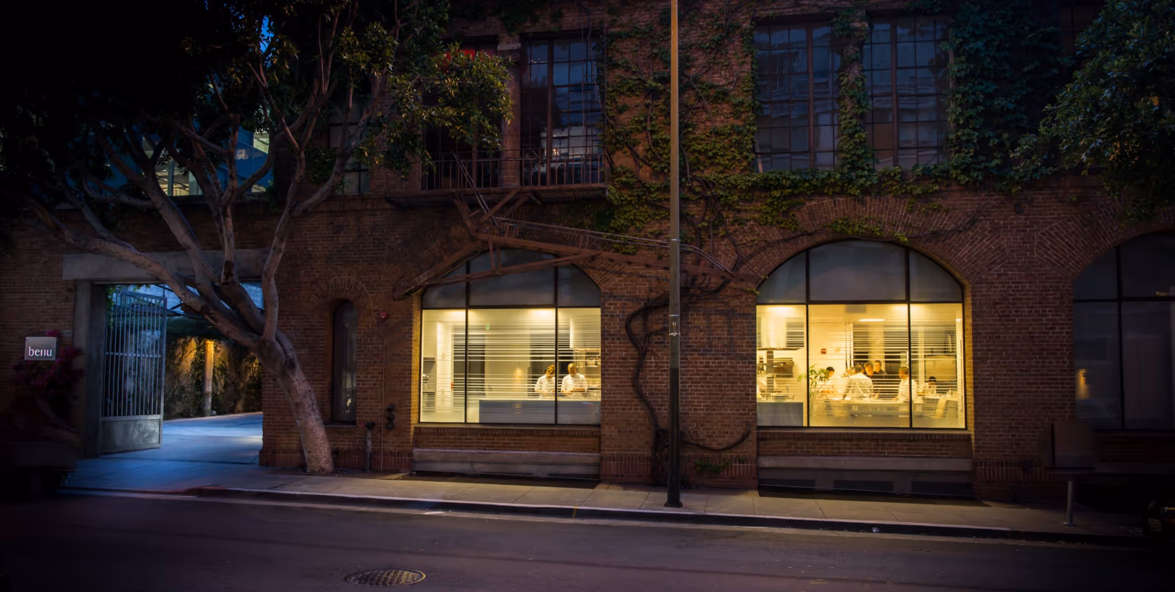 Night view of a brick building with large windows showing chefs working inside a warmly lit kitchen.