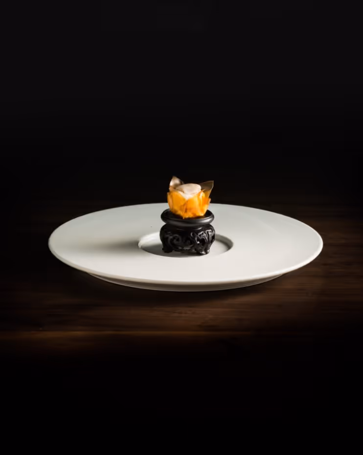 Small gourmet dessert with an orange translucent wrapper and cream topping served on a decorative black pedestal inside a large white plate.