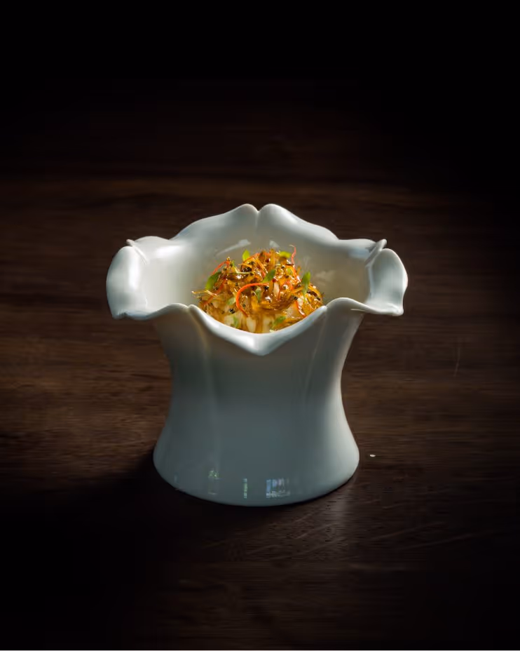 Elegant white flower-shaped ceramic bowl with a gourmet dish topped with finely sliced herbs and spices on a dark wooden surface.