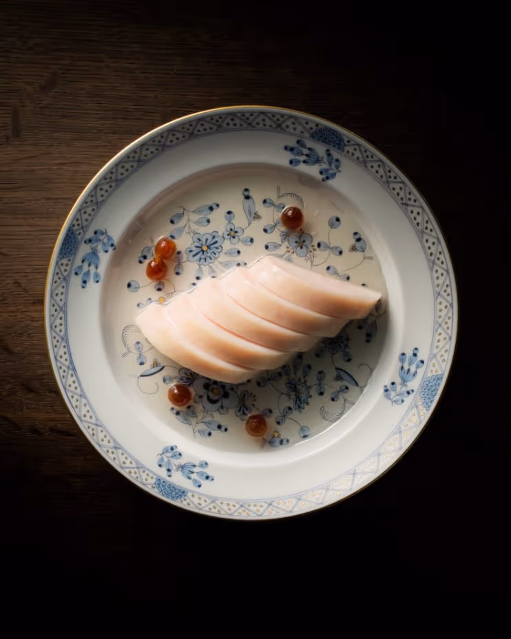 Sliced pale pink sashimi-style fish arranged in a curved row on a white plate with blue floral patterns, garnished with small red spheres.