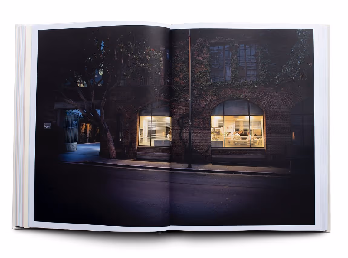 Open book showing a nighttime street view of a brick building with warmly lit windows and visible interior activity.