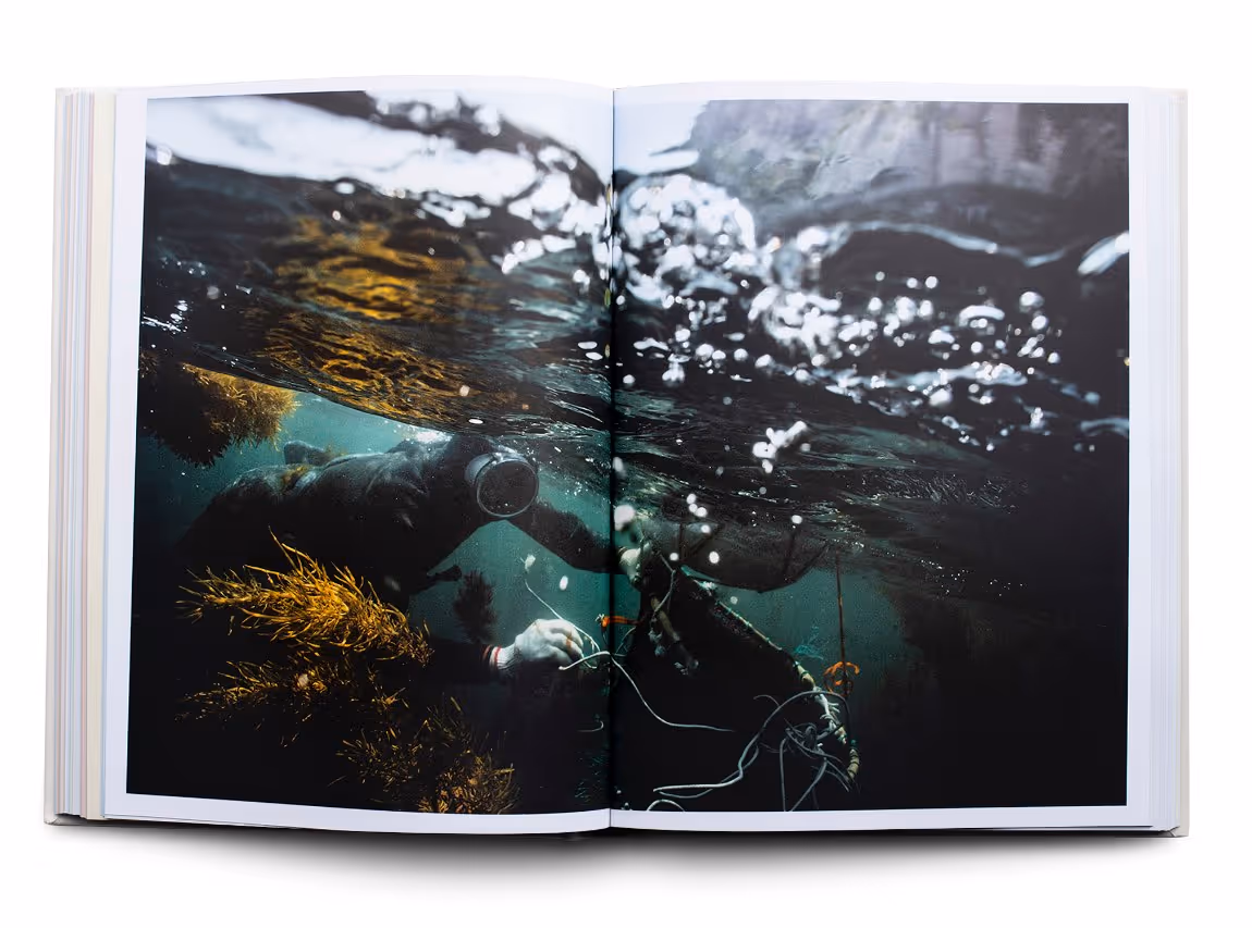 Open book showing a two-page underwater photograph of a diver in a wetsuit working among seaweed and ropes with water bubbles above.