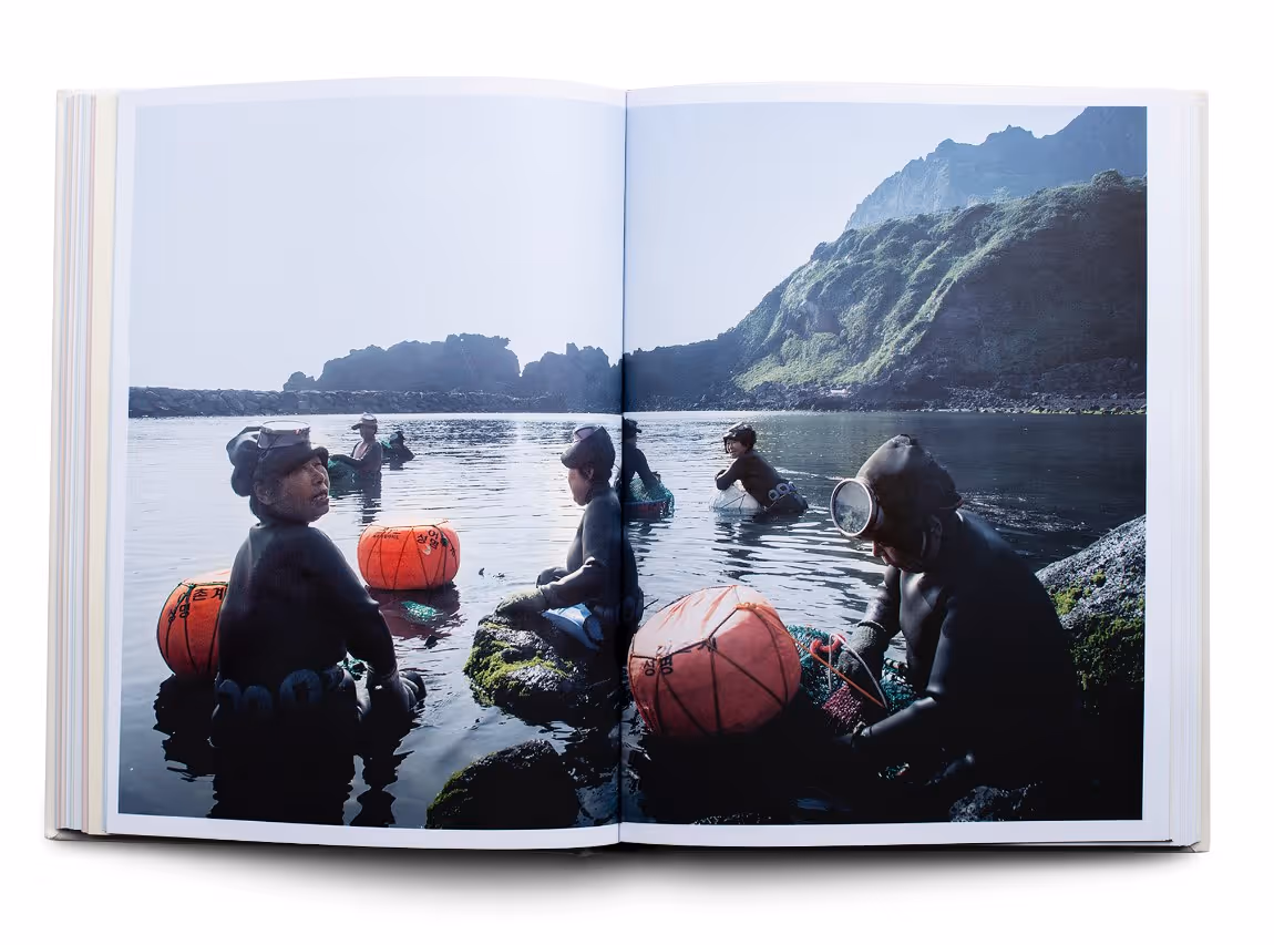 Open book showing a photograph of haenyeo divers in wetsuits with goggles and orange flotation buoys in coastal waters near rocky cliffs.