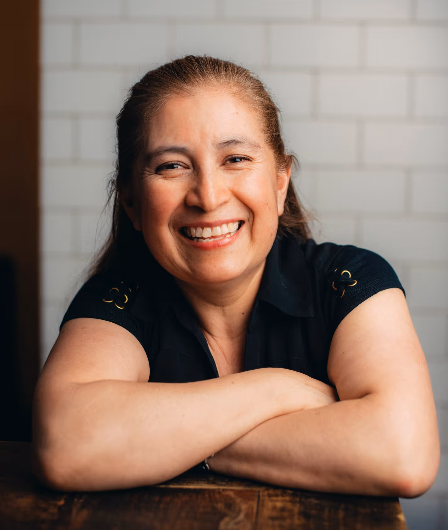 Smiling middle-aged woman in black shirt leaning on wooden table with folded arms.