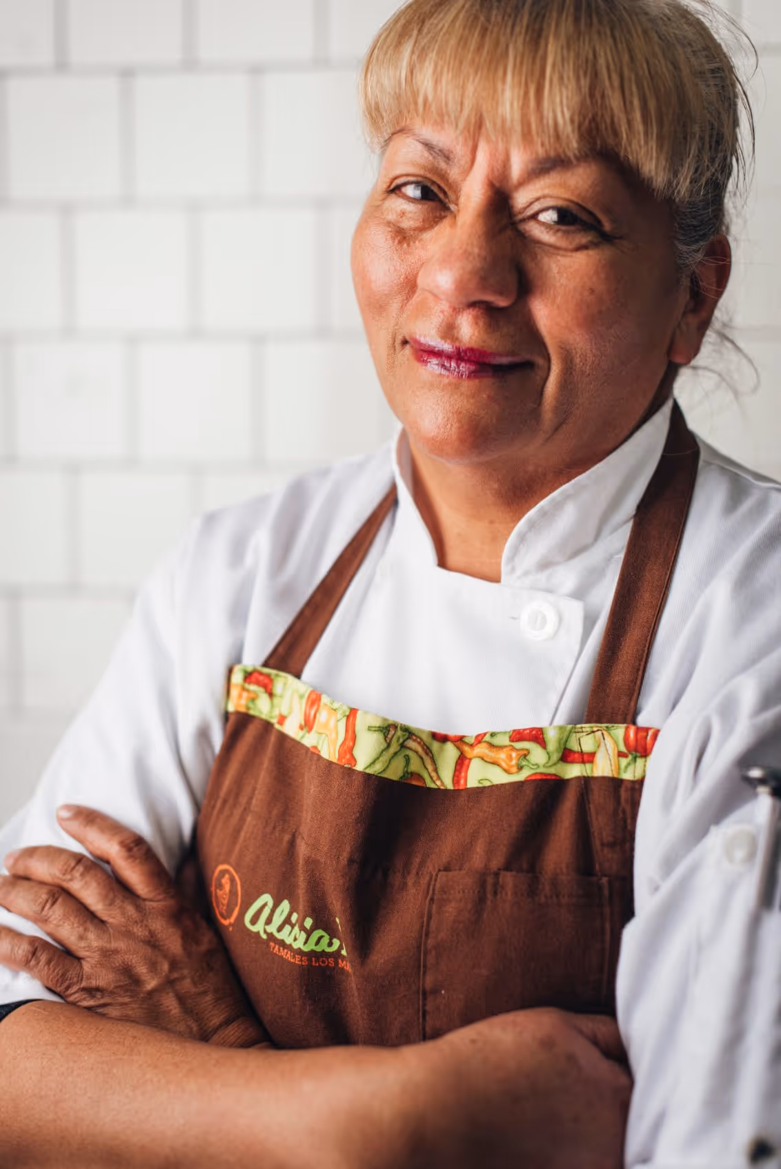 Smiling woman chef wearing a white coat and a brown apron with colorful trim, standing with arms crossed in a kitchen.