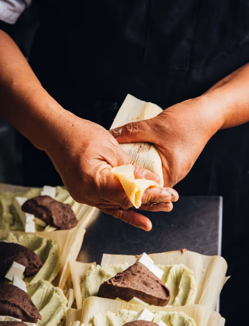 Person wrapping a tamale in corn husks with trays of tamales filled with masa and a dark-colored filling.