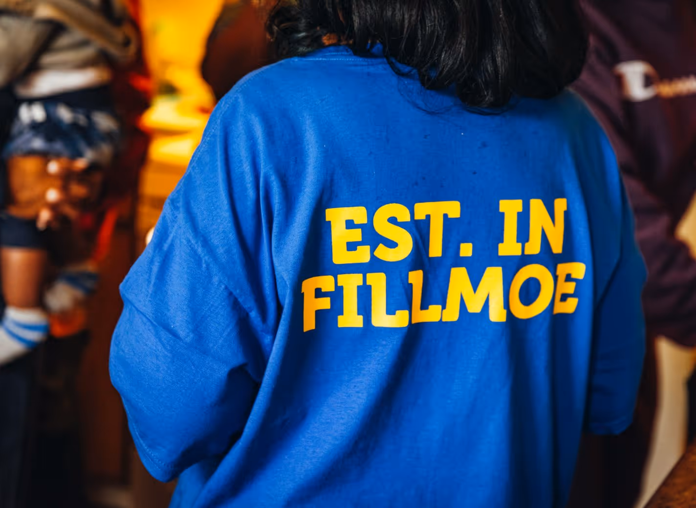 Person wearing a blue shirt with yellow text that reads 'EST. IN FILLMOE' on the back.