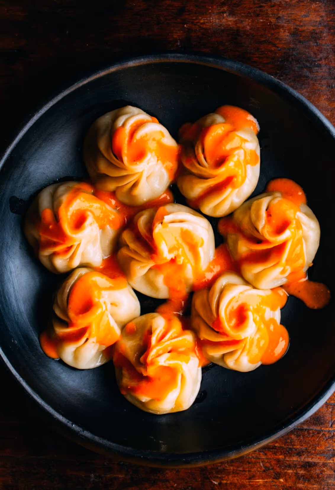 Seven steamed dumplings topped with a bright orange sauce on a black plate.