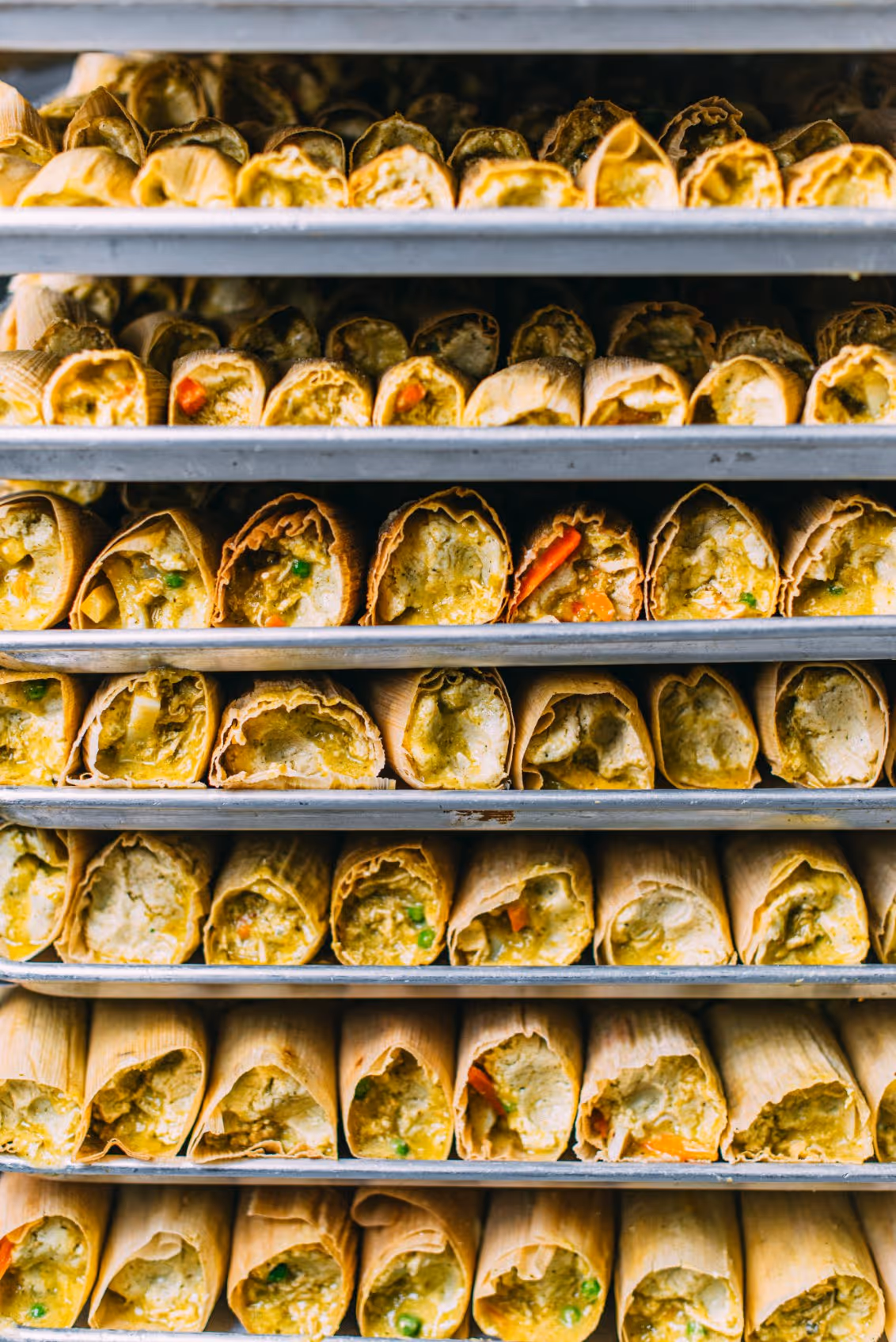Rows of uncooked tamales filled with seasoned dough and vegetables stacked on baking trays.