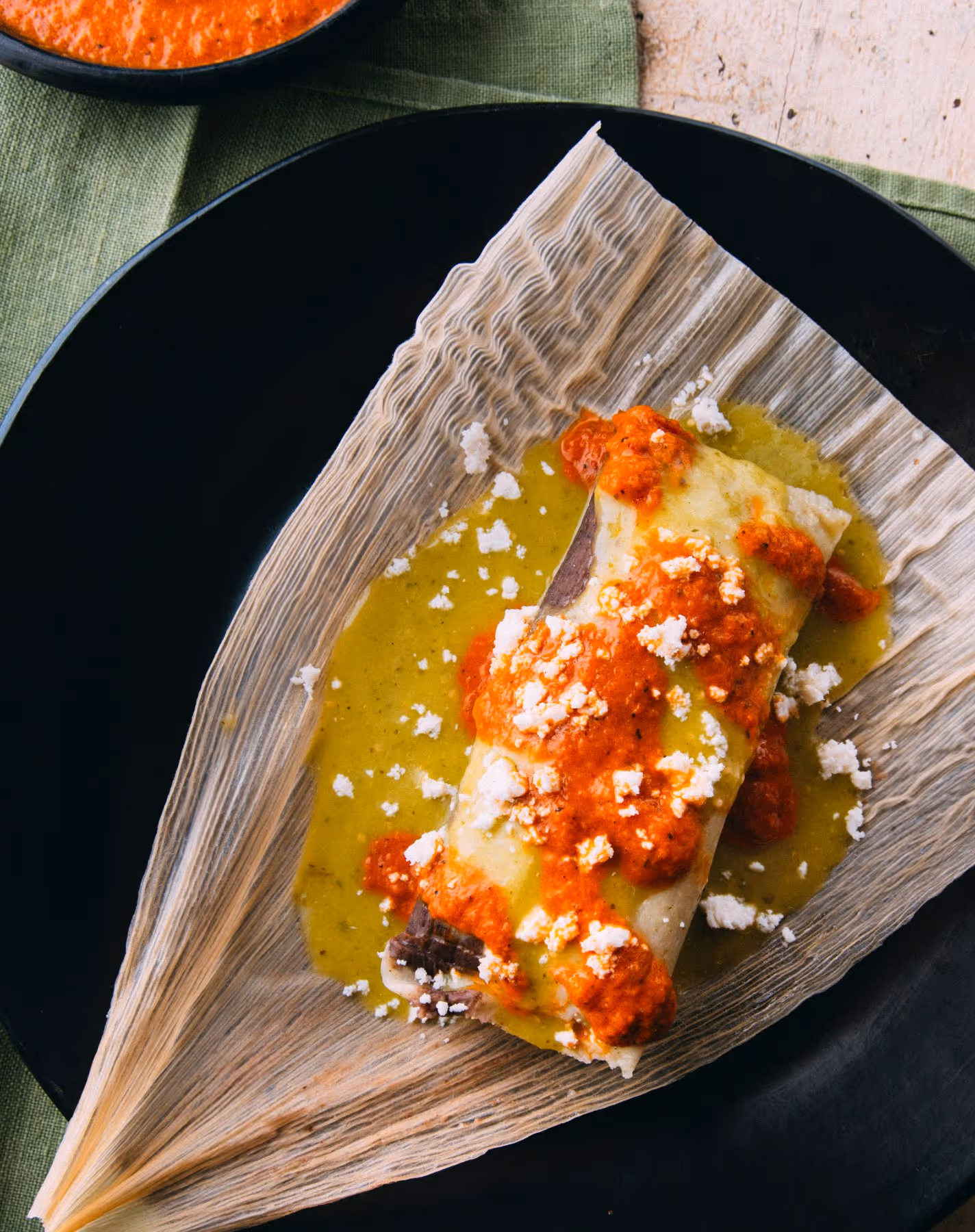 A tamale on a dried corn husk, topped with green and red sauce and crumbled cheese, served on a black plate.
