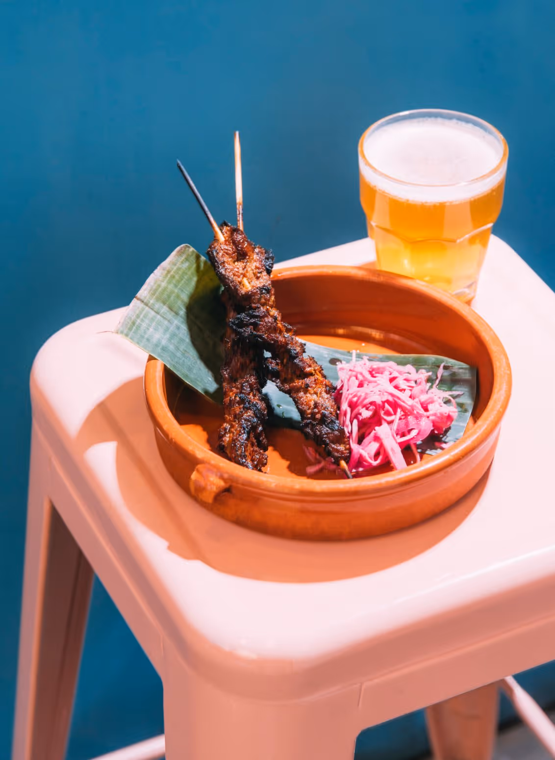 Two grilled meat skewers on a banana leaf with pickled red cabbage in a clay dish beside a glass of beer on a white stool.
