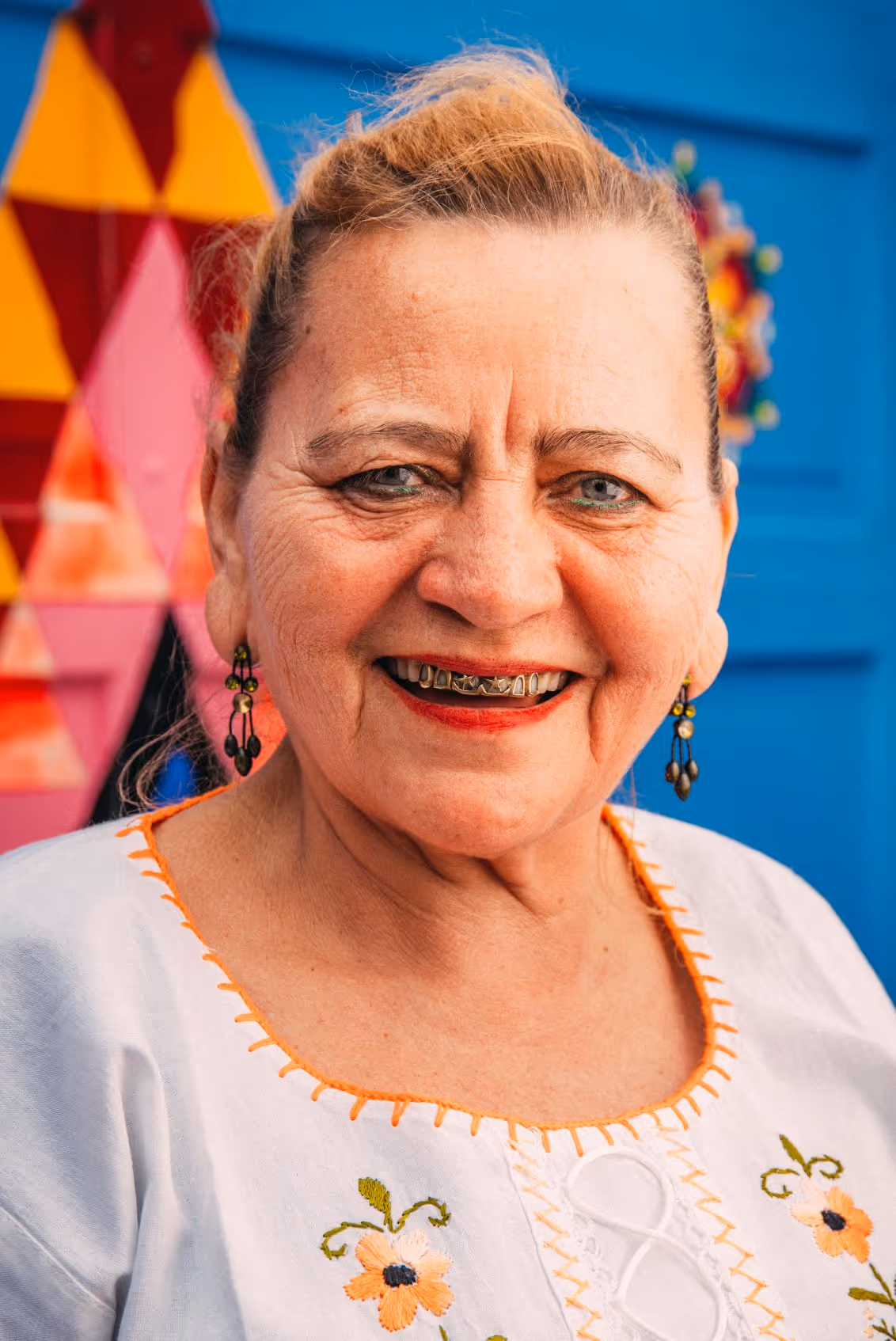 Smiling older woman wearing a white embroidered blouse and decorative grill teeth, standing in front of a colorful background.