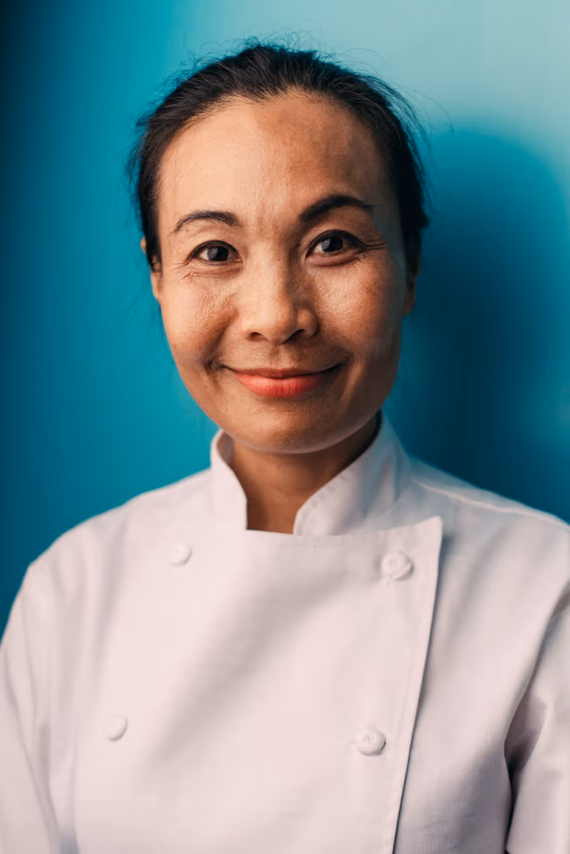 Smiling female chef wearing a white uniform against a turquoise background.