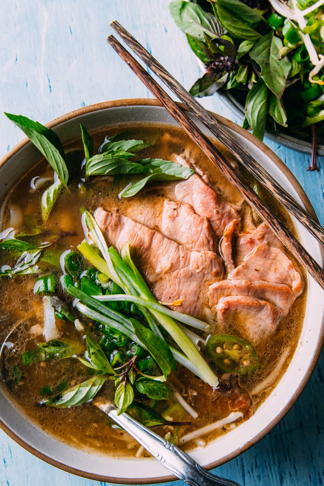 Bowl of pho with sliced beef, green onions, basil leaves, jalapeño slices, and chopsticks resting on the edge.