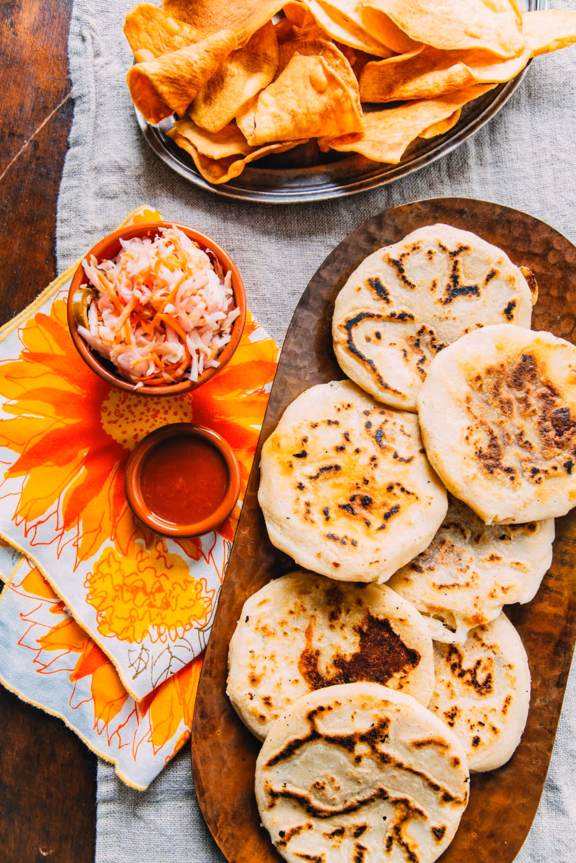 Plate of six grilled pupusas served with cabbage slaw and red salsa beside a plate of crispy tortilla chips.