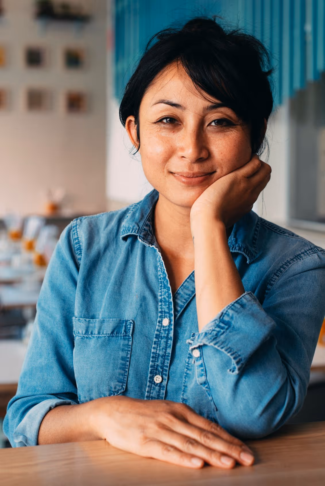 Smiling woman with dark hair wearing a denim shirt sitting at a table indoors with her hand resting on her face.