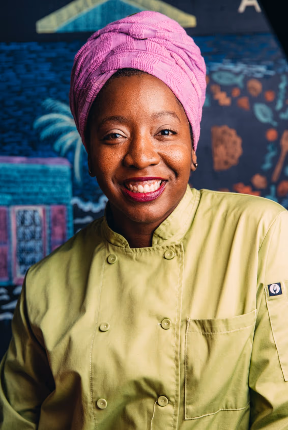 Smiling chef wearing a light green jacket and a bright pink headwrap with colorful mural background.