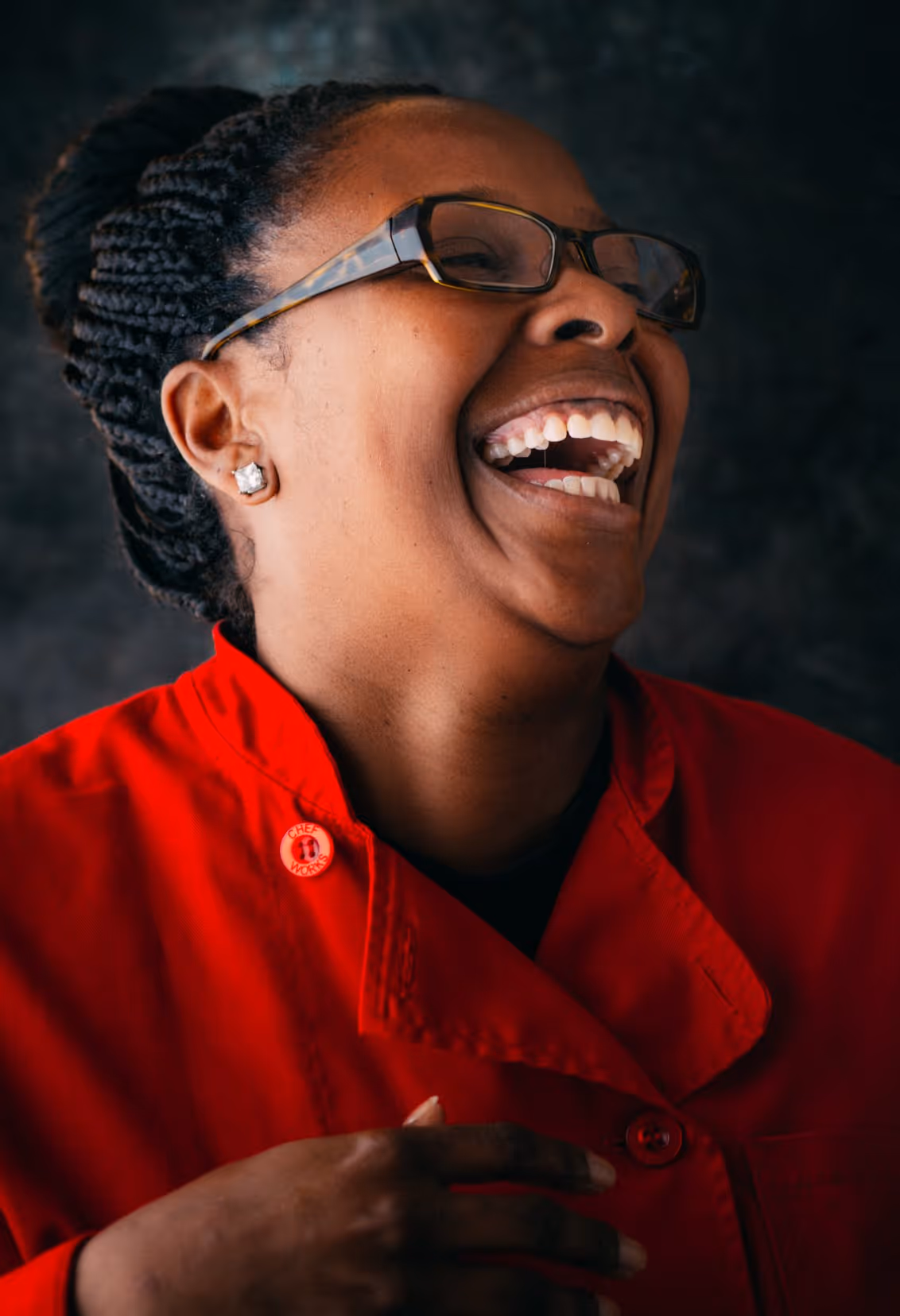 Woman with glasses and braided hair laughing joyfully while wearing a red chef coat.