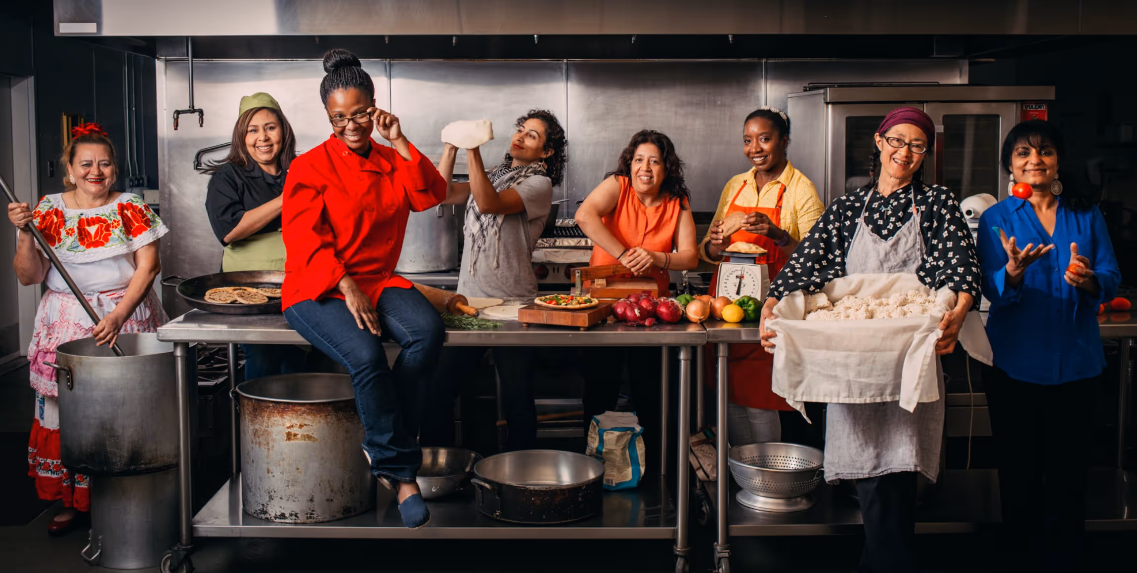Eight diverse women preparing traditional food together in a commercial kitchen, smiling and engaging with ingredients and cooking tools.