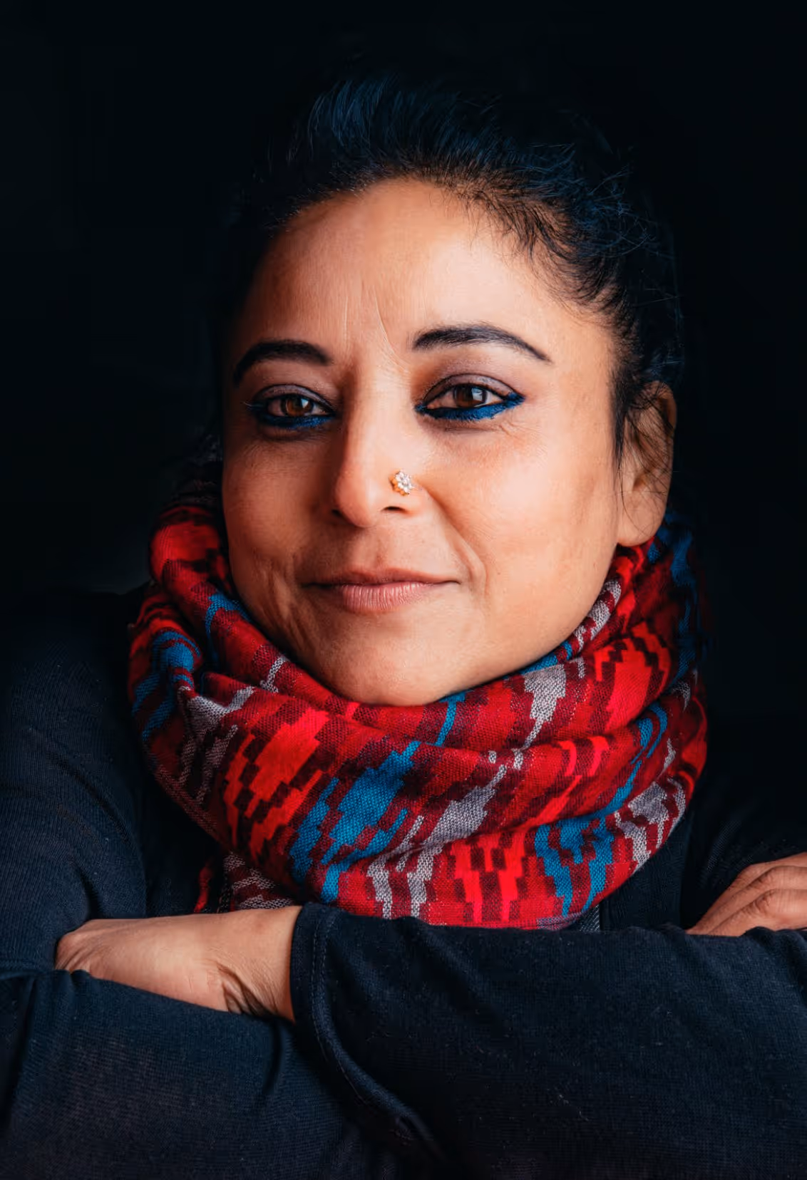 Woman with dark hair wrapped in a vibrant red, blue, and gray patterned scarf, wearing black clothing and blue eyeliner, smiling gently against a dark background.