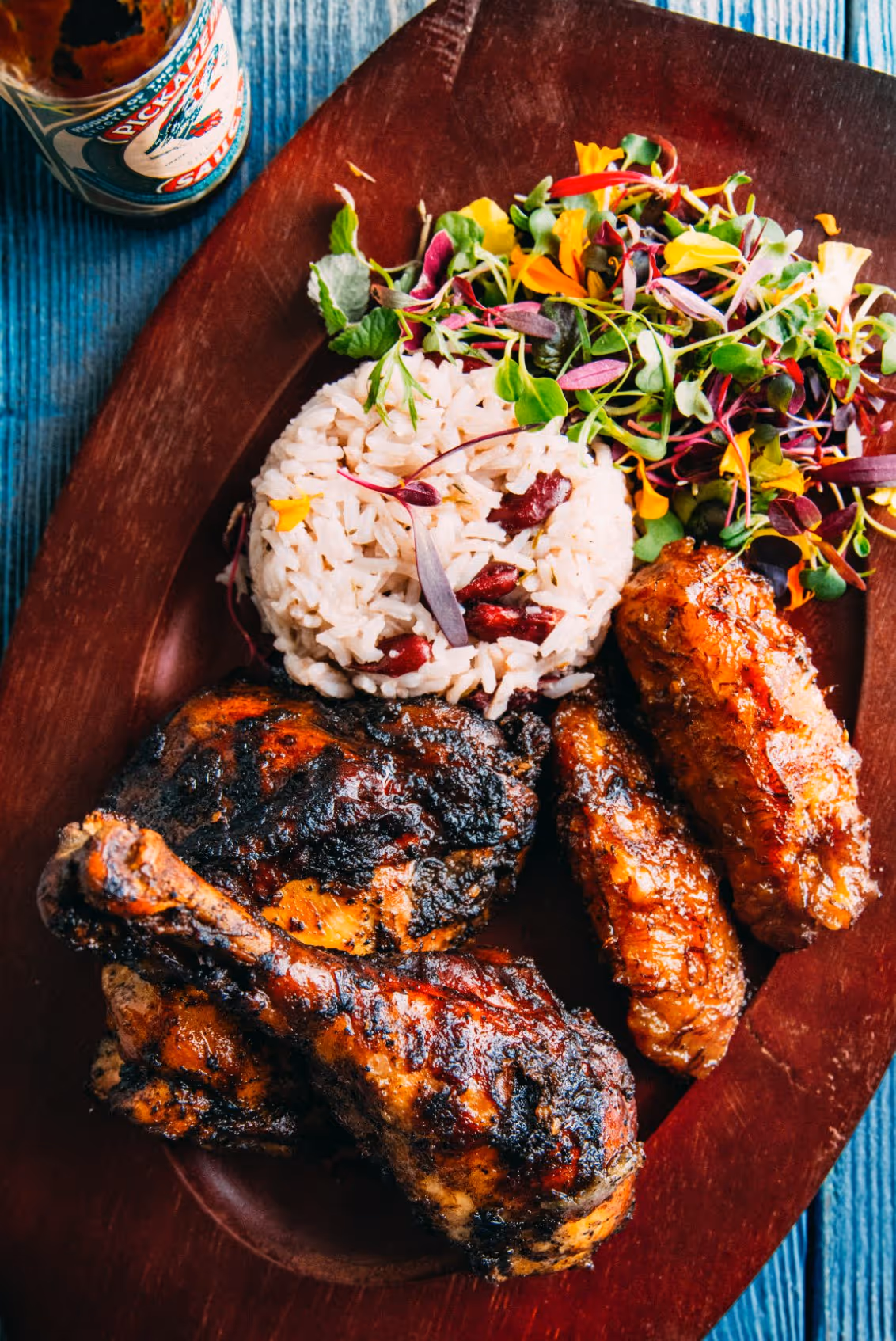 Grilled jerk chicken with rice and peas and a fresh herb salad on a wooden plate.