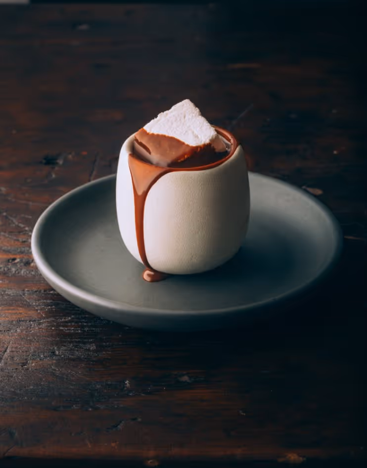 White ceramic cup with chocolate dripping down the side and a marshmallow dipped in chocolate inside, placed on a gray plate on a wooden surface.