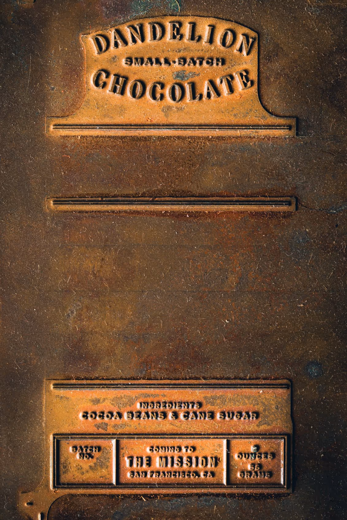 Rusty metal chocolate bar mold with embossed text reading 'Dandelion Small Batch Chocolate' and ingredients 'Cocoa Beans & Cane Sugar,' made in San Francisco.