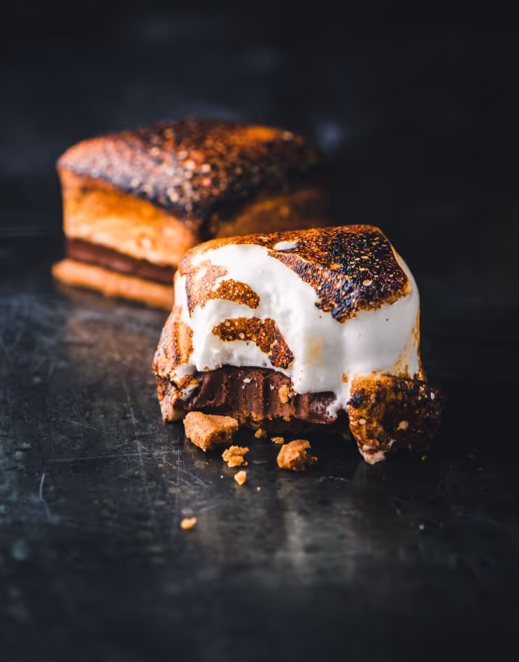 Close-up of two toasted marshmallow and chocolate s'mores on a dark surface, with one partially eaten showing melted chocolate.
