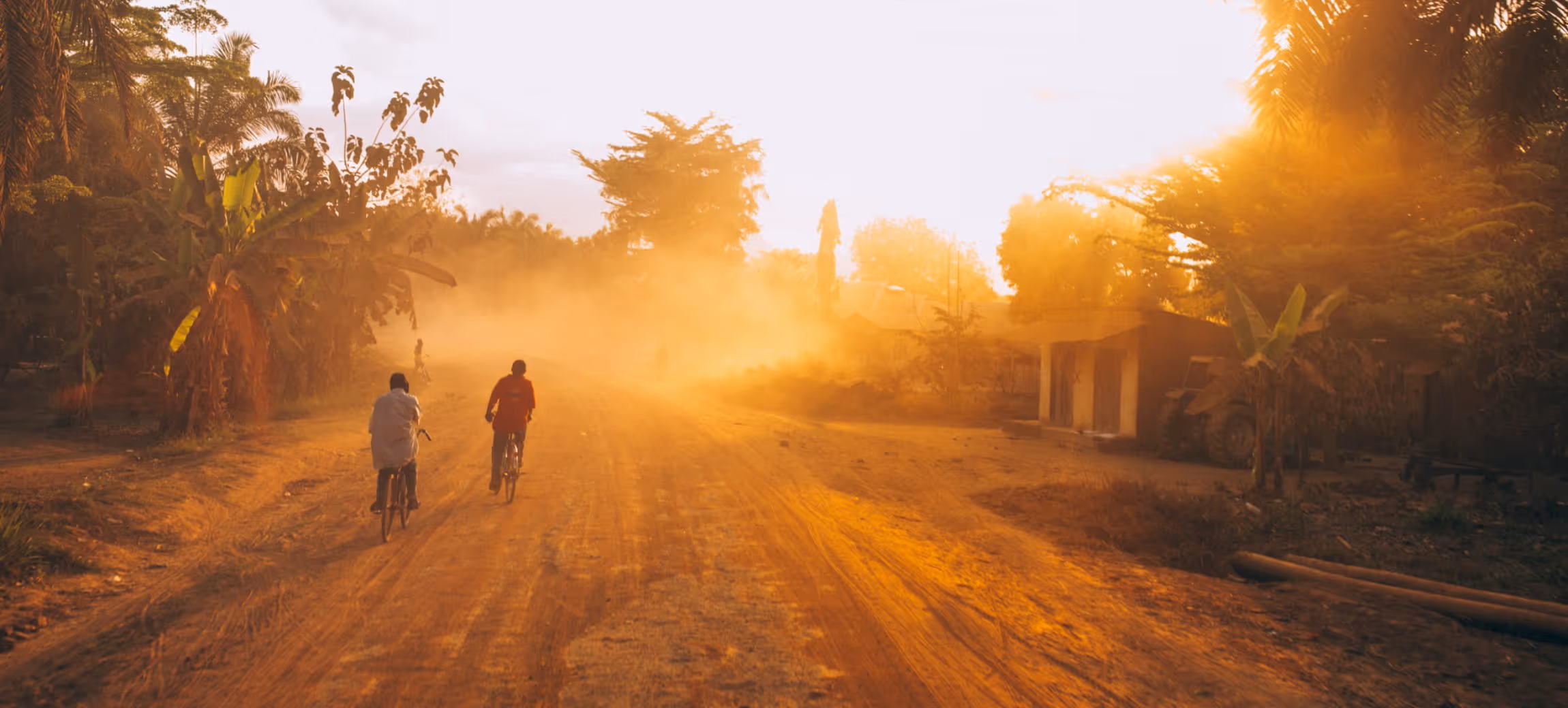Two people riding bicycles on a dusty road at sunset with lush greenery and houses on the sides.