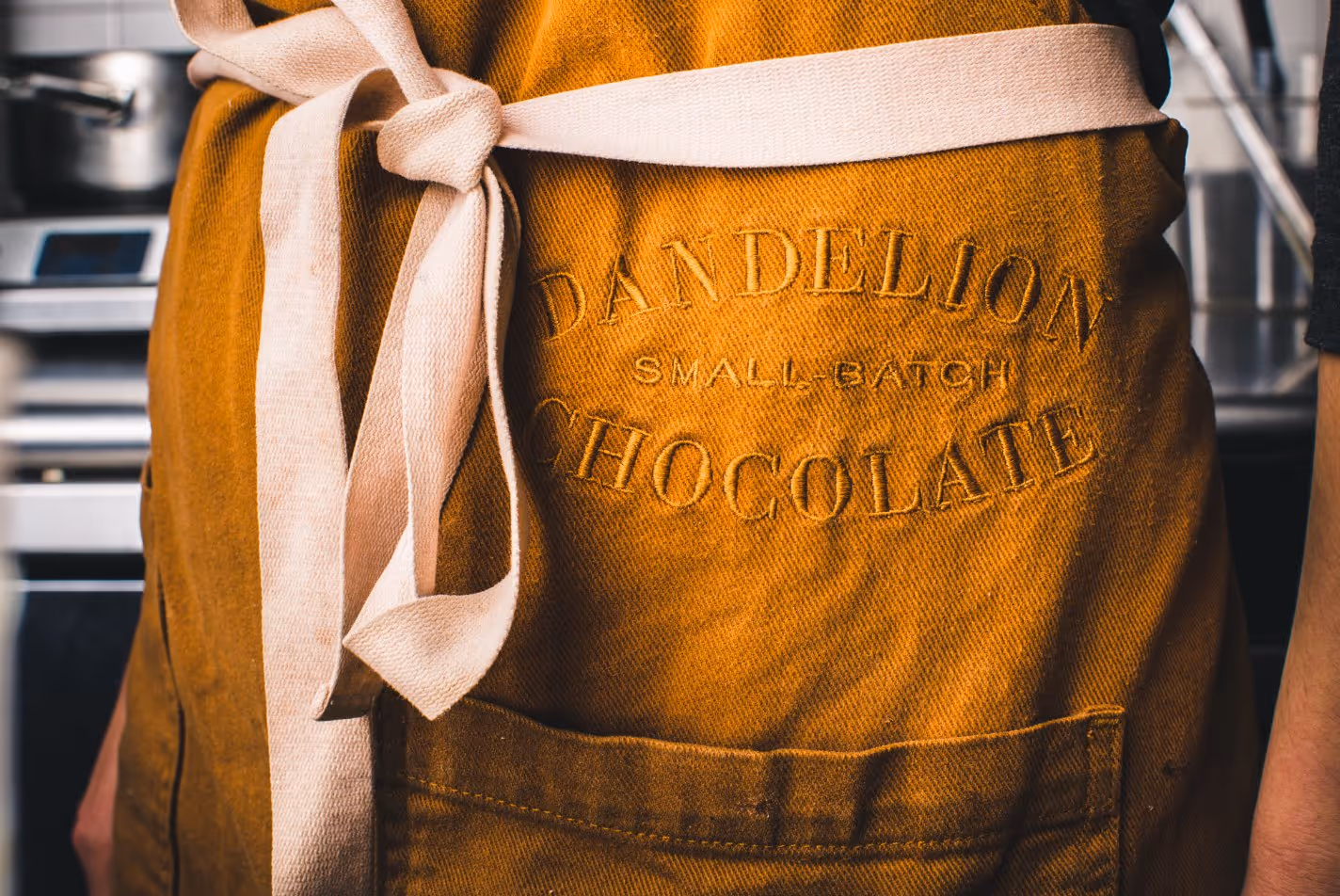 Close-up of a brown apron with a light beige tie, embroidered with the words 'Dandelion Small-Batch Chocolate'.