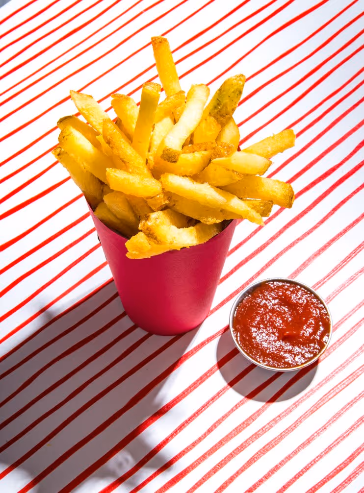 Golden French fries served in a red container with a side of red ketchup on a white surface with red diagonal stripes.